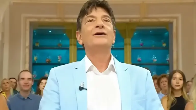 Un uomo in primo piano con giacca chiara, davanti a un gruppo di persone in un ambiente televisivo.