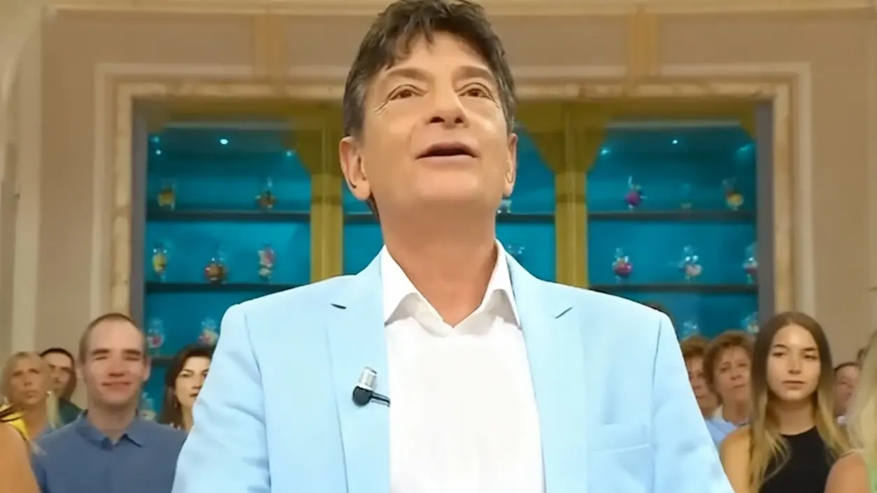 Un uomo in primo piano con giacca chiara, davanti a un gruppo di persone in un ambiente televisivo.